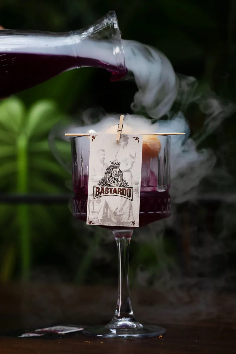 The Sparkling Mary Jane cocktail served with smoke in a tall glass with a BASTARDO tag, among the foliage