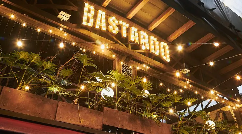 Illuminated BASTARDO sign above the entrance of the Provenza house, among string lights and plants